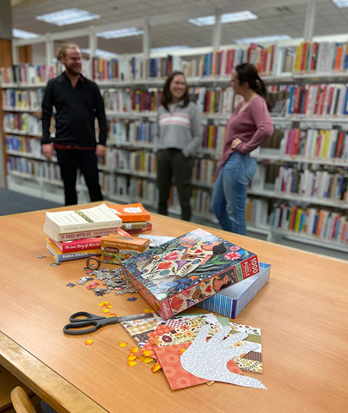Crafts, books and puzzles in the foreground. Three people talking to each other in the background.